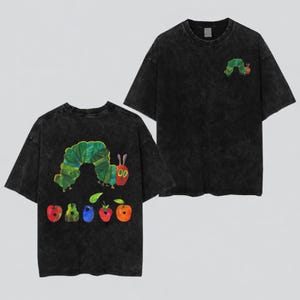 May include: A black acid-wash t-shirt featuring a small green caterpillar graphic on the front chest. The back of the shirt displays a large illustration of the same green caterpillar positioned above a row of five colorful fruits including an apple.