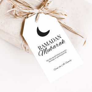 Op de afbeelding: Een wit cadeau-etiket met een zwarte halve maan en de tekst "RAMADAN Mubarak" in zwarte schrijfletters. De tekst eronder luidt "Moge deze maand u vrede, zegeningen en spirituele groei brengen. Van de familie Ali."