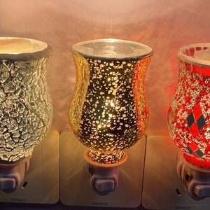 May include: Three decorative electric wax warmers with different mosaic patterns. The warmers are plugged into electrical outlets and have a light switch. The warmers are gold, silver, and red.