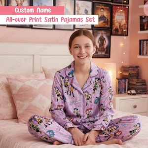 May include: A person wearing a lavender all-over print satin pajama set. The pajamas feature a repeating cartoon character design and the name "Taylor". The text "Custom Name" and "All-over Print Satin Pajamas Set" are visible.