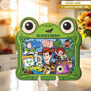 May include: A two-layered wooden sign shaped like a green frog with large eyes and a smiling mouth. The center features a colorful illustration of various animated toy characters, including a cowboy, space ranger, and cowgirl. The text OLIVIA'S ROOM is printed in green above the characters.