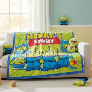 Może przedstawiać: Zielony i niebieski polarowy koc z kresk&oacute;wkowymi kosmitami z "Toy Story" z napisem "HENRY'S STORY". Koc jest narzucony na białą sofę, z pasującymi pluszowymi zabawkami i pudełkiem na zabawki w pobliżu. Koc to nadruk na zam&oacute;wienie.