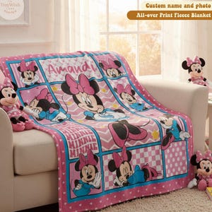 Może przedstawiać: R&oacute;żowy polar z Minnie Mouse, z imieniem "Amandi". Kocyk przedstawia wiele obraz&oacute;w Minnie Mouse i słowo "MINNIE" w wzorze kratki. Kocyk leży na białej sofie.