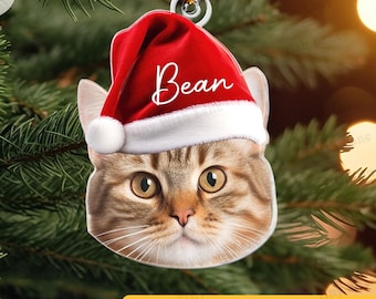 May include: A cat-shaped Christmas ornament with a red Santa hat that says "Bean" in white script. The ornament is hanging from a gold string against a backdrop of a Christmas tree. The text "Custom Name and Photo" is at the bottom.