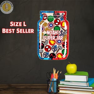 May include: A jar-shaped sign with a red, white, and blue border, filled with superhero logos. The text "NOAH'S SUPER JAR" is centered. The words "SIZE L" and "BEST SELLER" are displayed in red. A green apple rests on a stack of books.