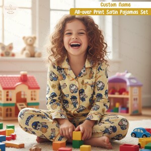 Puede incluir: Un ni&ntilde;o lleva un conjunto de pijama de sat&eacute;n amarillo con un estampado de personajes de dibujos animados. El pijama tiene una camisa abotonada y pantalones. El texto "Custom Name" y "All-over Print Satin Pajamas Set" son visibles.
