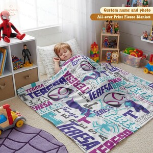 Może przedstawiać: Biały polarowy koc z powtarzającym się wzorem imienia "TERESA" w kolorze fioletowym, turkusowym i białym, wraz z grafiką pająka. Dziecko śpi pod kocem. Widoczny jest tekst "Custom name and photo All-over Print Fleece Blanket".