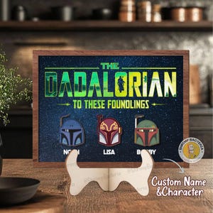 May include: A wooden framed sign with the text "THE DADALORIAN TO THESE FOUNDLINGS". The sign features three helmet designs with the names "NOI", "LISA", and "BBY". The sign is displayed on a wooden easel.