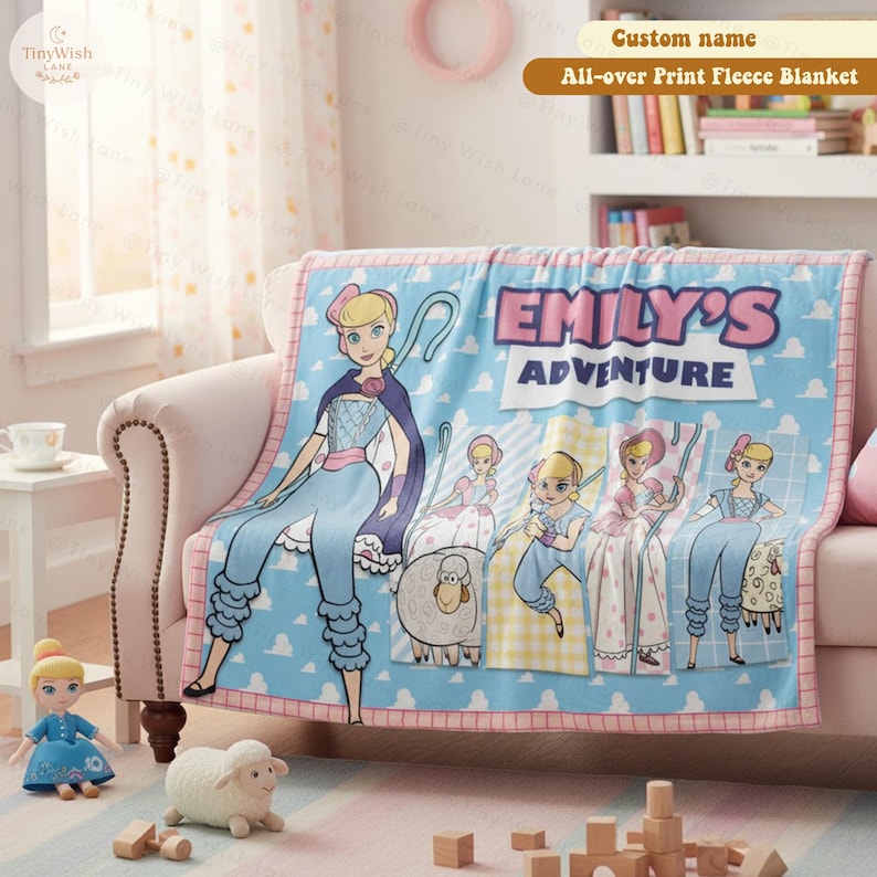 Może przedstawiać: Jasnoniebieski polarowy koc z r&oacute;żową obw&oacute;dką z postacią z kresk&oacute;wki. Koc ma napis "EMILY'S ADVENTURE" i "Custom name All-over Print Fleece Blanket". Koc jest narzucony na r&oacute;żową sofę.