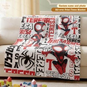 Może przedstawiać: Biały koc z polaru z powtarzającym się wzorem imienia "TERESA" w kolorze czerwonym i czarnym, wraz z ilustracjami superbohatera. Koc zawiera r&oacute;wnież dużą grafikę pająka.