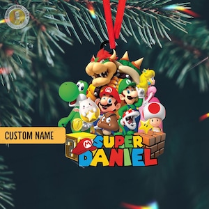 May include: A Christmas tree ornament featuring Super Mario characters. The ornament has a red hanging ribbon and the text "SUPER DANIEL" in bright colours. The background is a dark green, with a Christmas tree visible.
