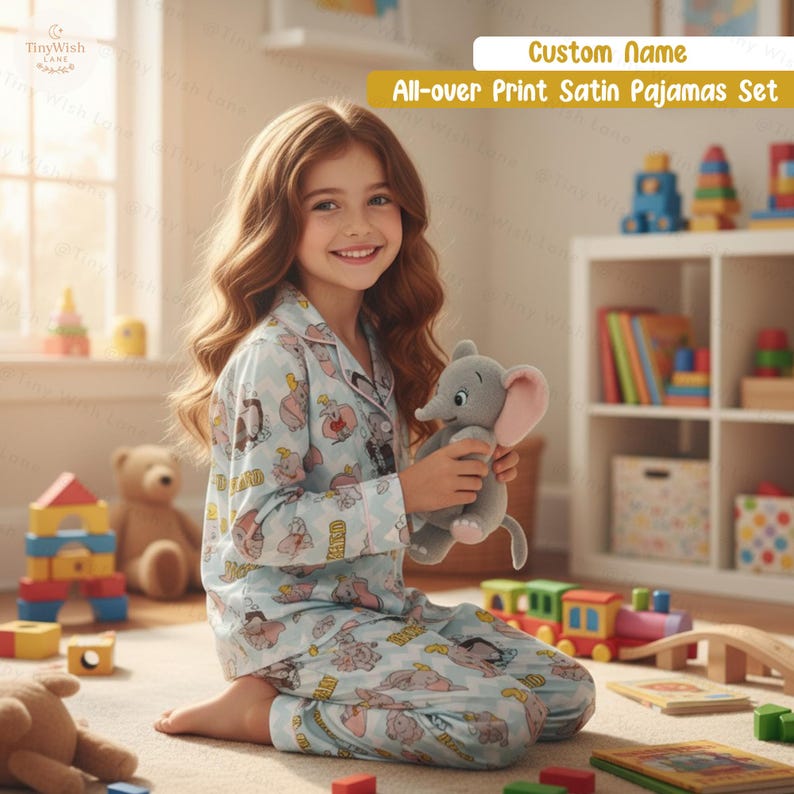 May include: A young person wearing a light blue all-over print satin pajama set, holding a grey elephant plush toy. The pajamas feature a cartoon design. The text on the image says "Custom Name" and "All-over Print Satin Pajamas Set".