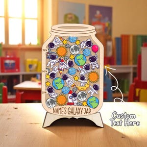 Puede incluir: Una pieza decorativa de madera con forma de tarro con temática espacial. El tarro está lleno de astronautas, planetas, cohetes y estrellas. El texto "NAME'S GALAXY JAR" está en la parte inferior. Montado sobre un soporte de madera. También se ve el texto "Custom Text Here".
