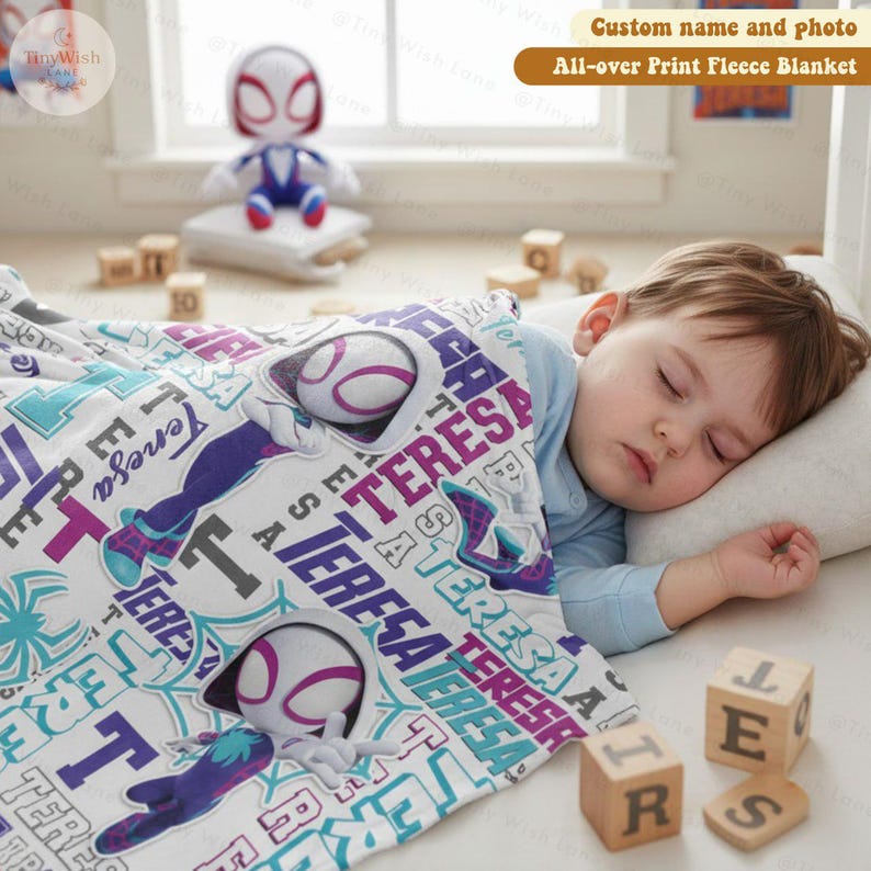 Może przedstawiać: Biały polarowy koc z powtarzającym się wzorem imienia "TERESA" w kolorze fioletowym i turkusowym, wraz z ilustracjami superbohatera. Koc przykrywa śpiące dziecko. Pasująca lalka znajduje się w tle. Widoczny jest tekst "Custom name and photo All-over Print Fleece Blanket".