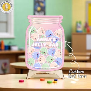 Puede incluir: Un letrero de madera rosa en forma de tarro con el texto "Anna's Jelly Jar" y lleno de gominolas de dibujos animados. El letrero está sobre un soporte de madera. Caramelos de colores están esparcidos sobre la mesa.