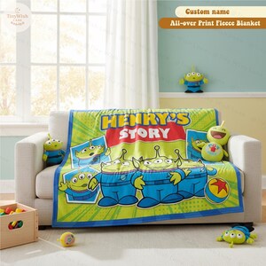 May include: A green and blue fleece blanket featuring cartoon aliens from Toy Story, with the text "HENRY'S STORY" in red and yellow. The blanket is draped over a white sofa, with matching plush toys and a toy box nearby.
