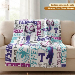 Może przedstawiać: Biały koc z polaru z nadrukiem na całej powierzchni przedstawiającym superbohaterkę. Koc zawiera imię "TERESA" w r&oacute;żnych czcionkach i kolorach, a także obrazy superbohater&oacute;w. Koc jest prezentowany na beżowej sofie.