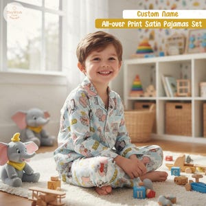 May include: A light blue satin pajama set with an all-over elephant cartoon print. The set includes a button-down shirt and trousers. A smiling child is wearing the pajamas in the image.