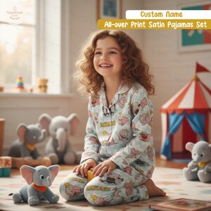 May include: A light blue satin pajama set featuring a pattern of pink elephants. The pyjamas have a button-down top and matching trousers. The text "Custom Name" and "All-over Print Satin Pajamas Set" are visible. A young person is wearing the pyjamas.