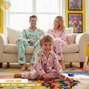 Op de afbeelding: Een gezin draagt bijpassende satijnen pyjama's met tekenfilmprints. De pyjama's zijn lichtblauw en roze. Het kind op de voorgrond speelt met kleurrijke bouwstenen. De tekst op de afbeelding luidt "Custom Name All-over Print Satin Pajamas Set."