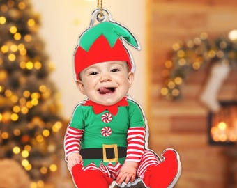 May include: A Christmas ornament featuring a smiling baby dressed as an elf. The baby wears a green and red elf costume with a matching hat and shoes. The ornament is designed to hang and is perfect for holiday decor.