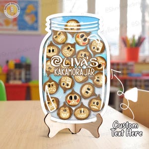 May include: A wooden jar-shaped sign with the text "Oliva's Kakamora Jar" and a collection of cartoonish coconut characters. The sign is propped up on a wooden stand. The background is a light blue sky.