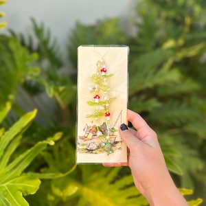 May include: A green frog charm hangs from a beaded necklace with green leaves and red beads. The necklace is displayed on a card with a whimsical illustration of a frog in a boat.