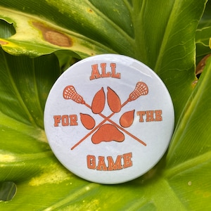 Könnte beinhalten: Ein weißer Knopf mit einem Pfotenabdruck und Lacrosse-Schlägern. Der Text "ALL FOR THE GAME" ist auf dem Knopf gedruckt.