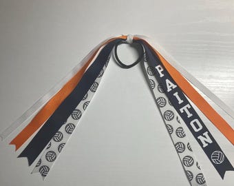 May include: A white, orange, and navy blue hair tie with a volleyball pattern and the name "PAITON" printed on one of the ribbons.