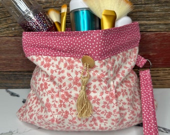 May include: A pink and white floral print makeup bag with a gold tassel and a pink polka dot strap. The bag is filled with makeup brushes, a bottle of blue liquid, and a bottle of clear liquid.