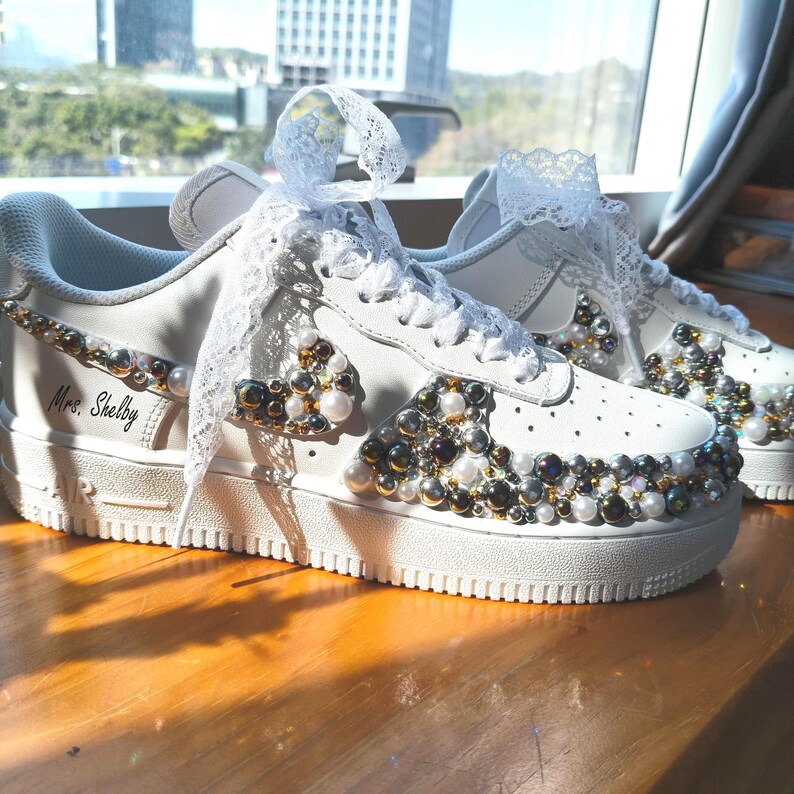 Puede incluir: Zapatillas blancas adornadas con perlas y cuentas iridiscentes. Los zapatos presentan detalles de encaje en los cordones y el logotipo de Nike. El texto "Mrs. Shelby" est&aacute; escrito en el lateral del zapato.