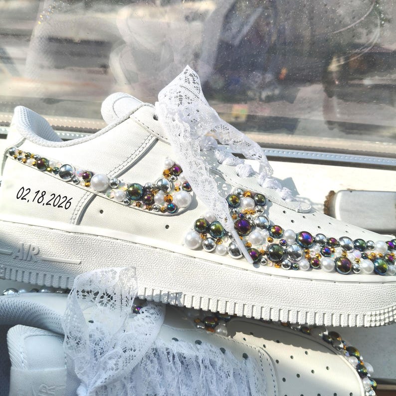 Puede incluir: Zapatillas blancas adornadas con perlas y gemas iridiscentes. Los zapatos presentan una superposici&oacute;n de encaje y la fecha "02.18.2026" impresa en el lateral. Las zapatillas tienen un dise&ntilde;o cl&aacute;sico con suela de goma.