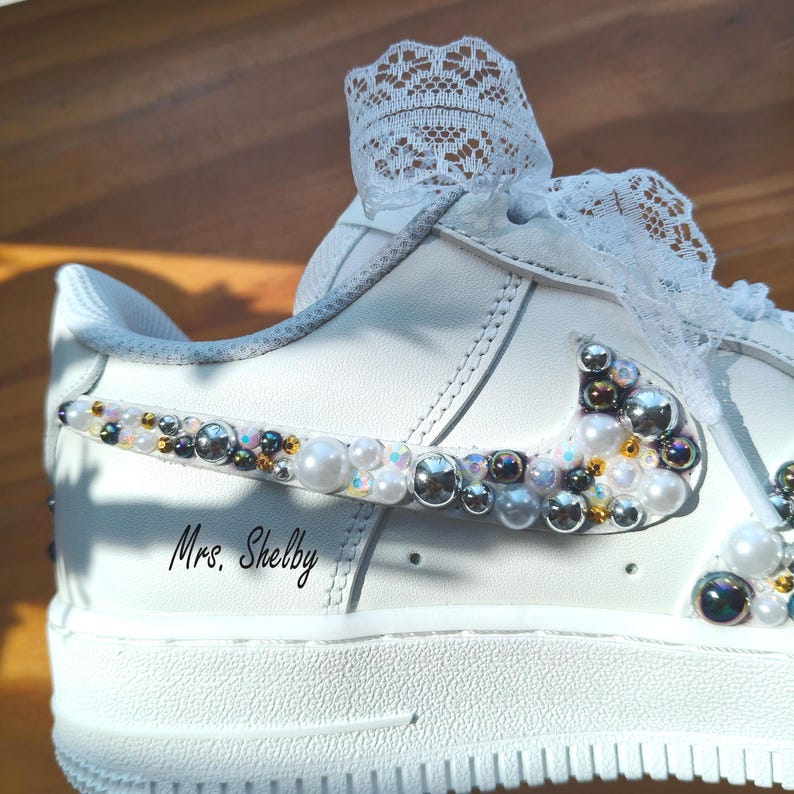 Puede incluir: Zapatilla blanca adornada con perlas y pedrer&iacute;a. El logo de Nike y el panel lateral est&aacute;n adornados con perlas y pedrer&iacute;a iridiscente de varios tama&ntilde;os. El texto "Mrs. Shelby" est&aacute; escrito en el lateral. Un detalle de encaje blanco est&aacute; en la parte superior.