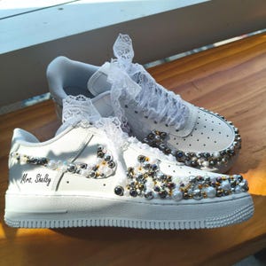Puede incluir: Zapatillas blancas adornadas con perlas y cristales. Los zapatos presentan detalles de encaje en los cordones y est&aacute;n personalizados con el nombre "Mrs. Shelby". Las suelas son blancas.