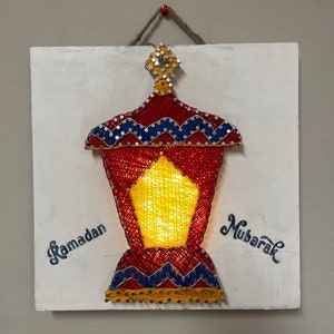 May include: A white wooden square with a string lantern made of yarn in red, blue, and yellow. The lantern has a light inside and the words "Ramadan" and "Mubarak" are written on the wood.
