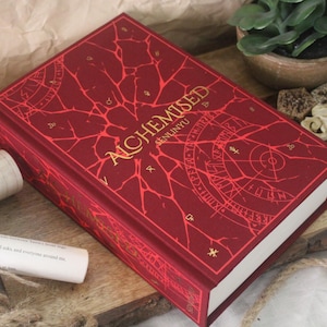 Op de afbeelding: Een hardcover boek getiteld Alchemised door Senlinyu met een dieprode kaft, gouden belettering en ingewikkelde rode barstpatronen. De voorkant toont cirkelvormige geometrische diagrammen en alchemistische symbolen in een ontwerp.