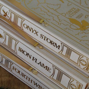 May include: A stack of four white books with gold accents and titles. The top book is titled "ONYX STORM" with a map on the cover. The second book is titled "IRON FLAME", the third book is titled "FOURTH WIN", and the bottom book's title is not visible.