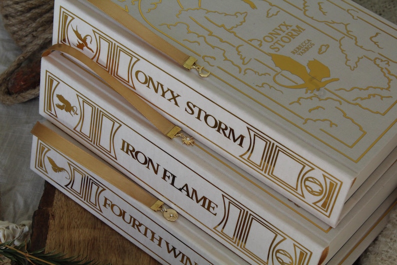 May include: A stack of four white books with gold accents and titles. The top book is titled "ONYX STORM" with a map on the cover. The second book is titled "IRON FLAME", the third book is titled "FOURTH WIN", and the bottom book's title is not visible.