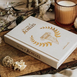 May include: A white book with gold lettering titled "The Song of Achilles" by Madeline Miller. The book is lying on a wooden surface with a sprig of white flowers and a rope coil.