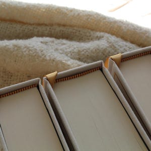 May include: Three white hardback books with gold edging. The books are stacked on top of each other.