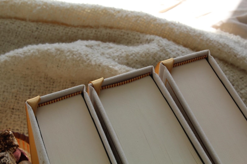 May include: Three white hardback books with gold edging. The books are stacked on top of each other.
