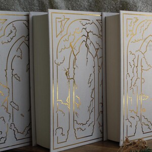 May include: Three white hardback books with gold foil designs on the covers. Each book features a different design of a tree with branches and leaves, framed by a gold arch.