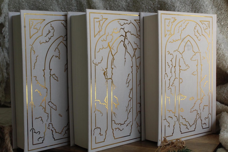 May include: Three white hardback books with gold foil designs on the covers. Each book features a different design of a tree with branches and leaves, framed by a gold arch.