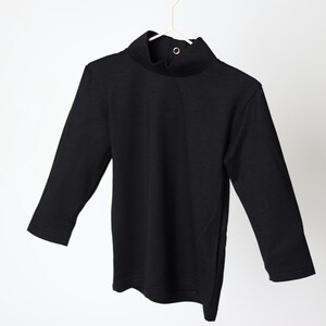 May include: A black long-sleeved shirt with a mock neck collar and a single button closure. The shirt is hanging on a white hanger against a white background. The garment is made of a soft, opaque fabric.