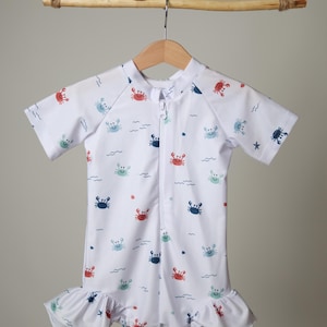 May include: White short-sleeved swimsuit with a crab pattern. The one-piece suit has a zipper down the front and a ruffled hem. The crabs are red, blue, and green, with wavy lines and stars scattered across the fabric.