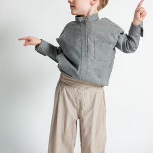 Puede incluir: Un ni&ntilde;o con una camisa gris de manga larga con un bolsillo en el pecho y pantalones beige de pernera ancha. La camisa tiene cuello mandar&iacute;n. El ni&ntilde;o est&aacute; se&ntilde;alando con ambas manos.