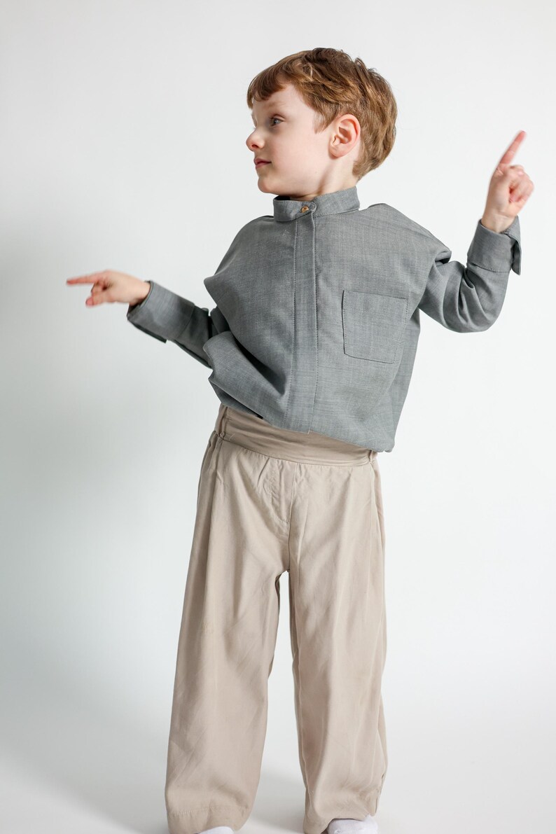 Puede incluir: Un ni&ntilde;o con una camisa gris de manga larga con un bolsillo en el pecho y pantalones beige de pernera ancha. La camisa tiene cuello mandar&iacute;n. El ni&ntilde;o est&aacute; se&ntilde;alando con ambas manos.