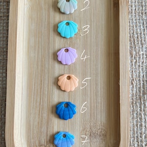 Può includere: Otto ciondoli a forma di conchiglia di diversi colori con un foro per la corda. I ciondoli sono disposti in fila su un vassoio di legno. I ciondoli sono dei seguenti colori: azzurro, bianco, blu, viola, arancione, blu scuro, lilla e blu scuro con vortici bianchi.
