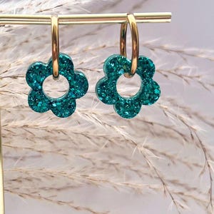 May include: A pair of gold hoop earrings with teal glitter flower charms. The earrings are displayed on a stand with a blurred background.