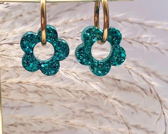 Könnte beinhalten: Ein Paar goldene Creolen mit tealfarbenen Glitzerblumen-Anhängern. Die Ohrringe werden auf einem Ständer mit verschwommenem Hintergrund präsentiert.